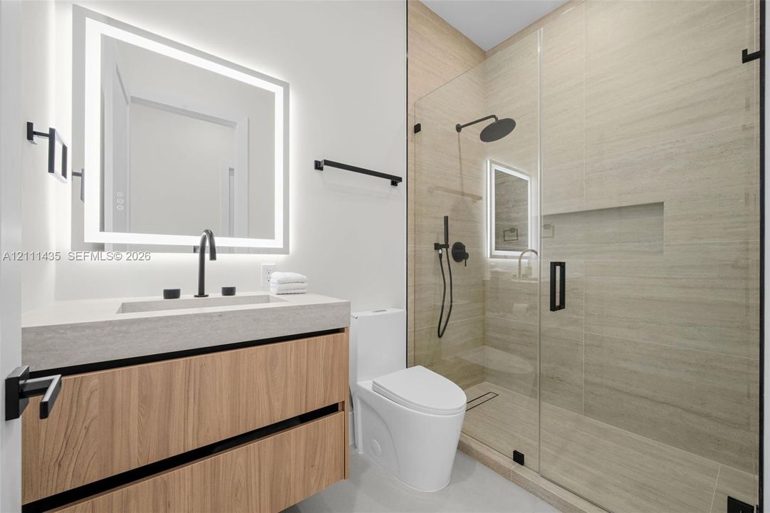 Floating wood-finish vanity with a rectangular sink, integrated LED-backlit mirror, walk-in shower with a glass enclosure and built-in recessed shelving, and matte black fixtures