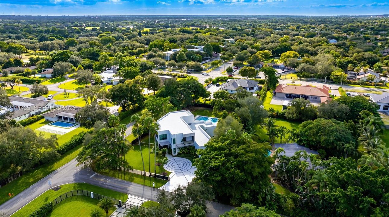 Aerial perspective of a multi-level residence featuring a circular patterned paved motor court, expansive manicured lawn, perimeter landscaping, and a rectangular swimming pool