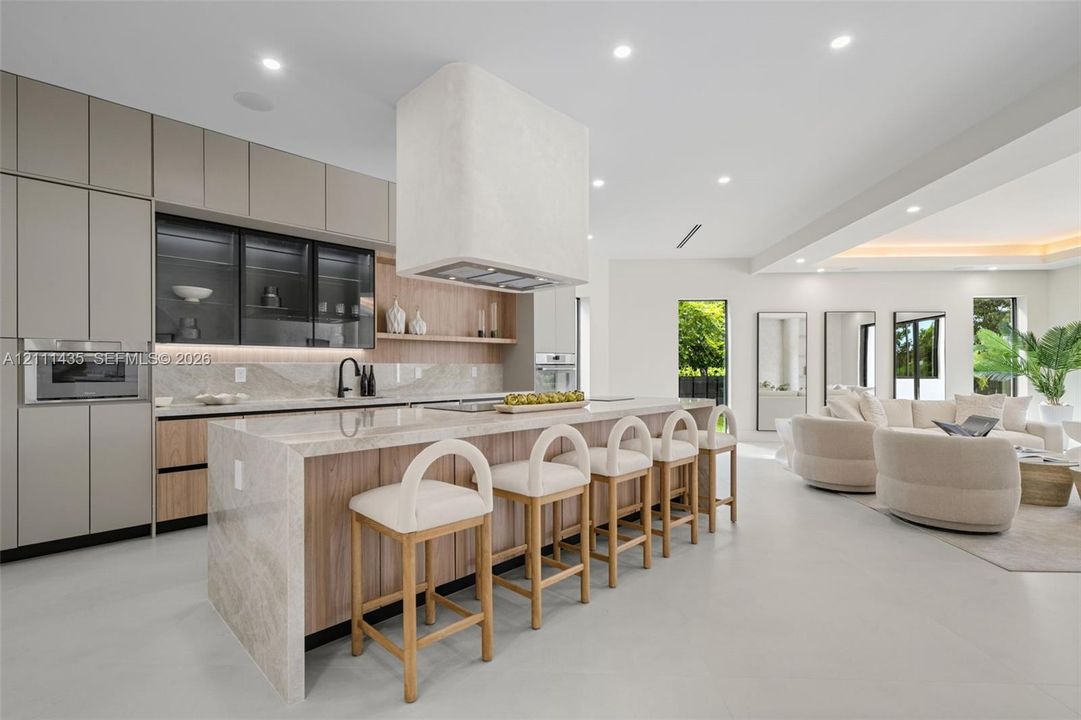 Modern kitchen featuring matte-finish cabinetry, a waterfall-edge stone island, integrated oven and microwave, recessed lighting, and a custom plaster range hood