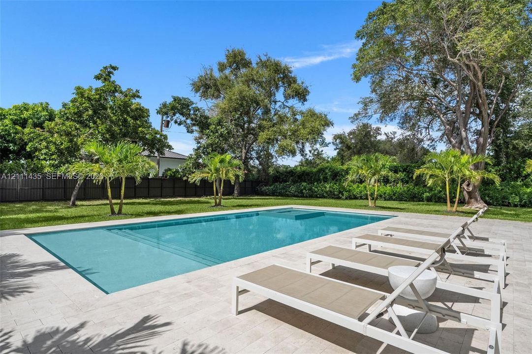 Rectangular swimming pool with integrated sun shelf and spa, surrounded by stone-finish paver decking, expansive grass lawn, mature trees, and privacy fencing