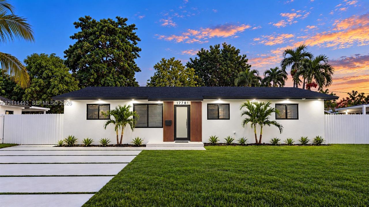 Single-story exterior featuring white stucco finish, wood-accented entryway, dark-toned roof shingles, black-framed windows, and a paved driveway with turf-filled expansion joints