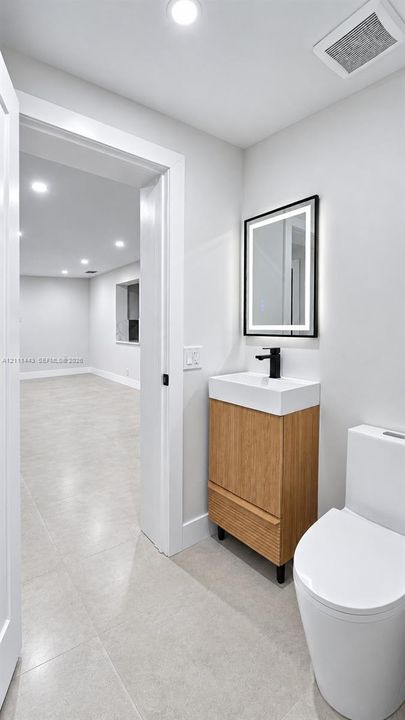 Modern vanity with wood-finish cabinetry, white basin, matte black faucet, and rectangular lighted mirror, complemented by ceramic tile flooring and a minimalist dual-flush toilet