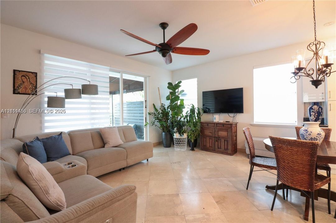 Neutral-toned stone flooring, five-blade ceiling fan, multi-arm iron chandelier, and glass sliding door featuring horizontal blinds