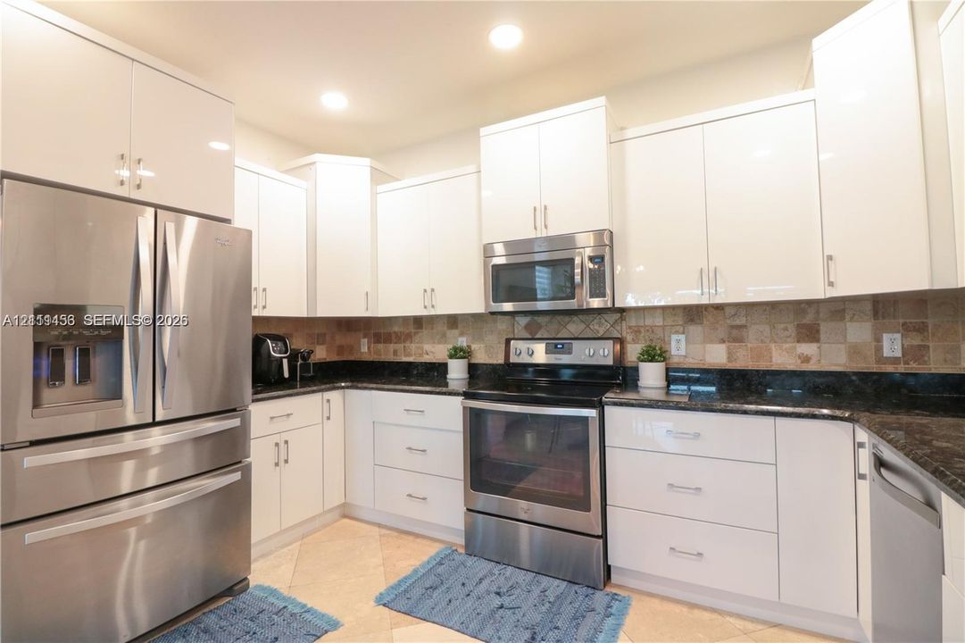 Kitchen featuring high-gloss white cabinetry, dark stone countertops, stainless steel appliances, and a decorative tile backsplash