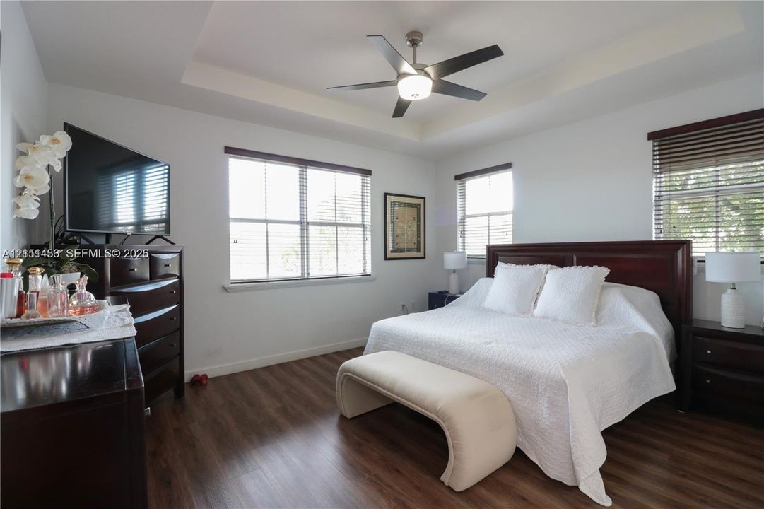 Tray ceiling with a central multi-blade ceiling fan, wood-finish flooring, and multiple windows with horizontal blinds