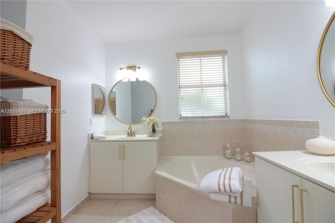 Bathroom featuring a corner soaking tub with tile surround, dual white vanity cabinets, round gold-rimmed mirrors, and light fixtures