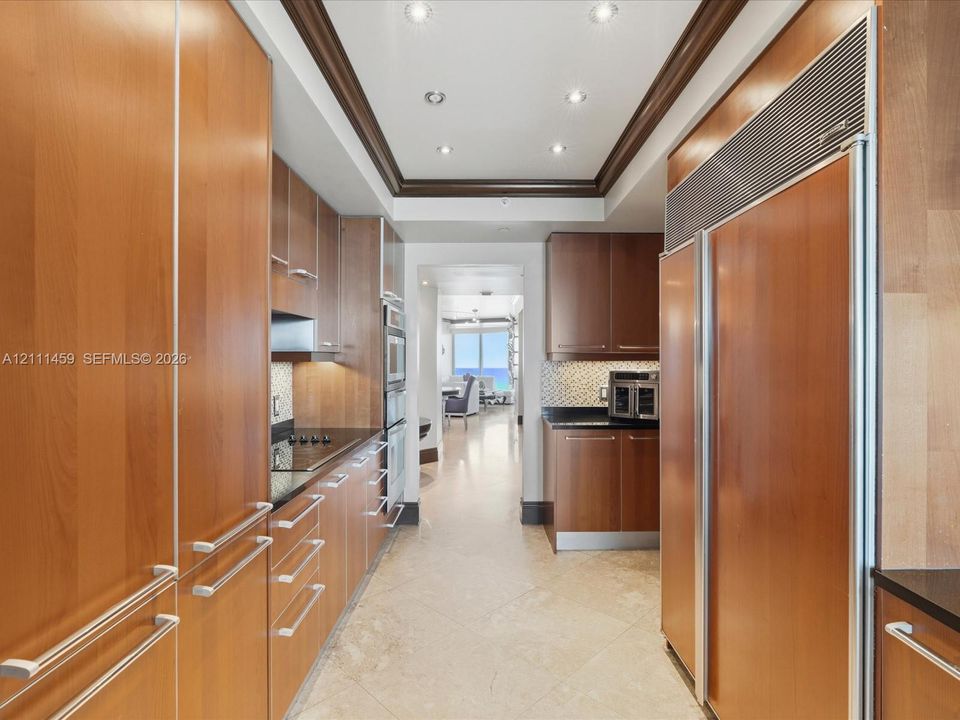 Galley kitchen featuring custom flat-panel wood cabinetry, integrated stainless steel refrigerator, black stone countertops, tile flooring, and recessed ceiling lighting with crown molding