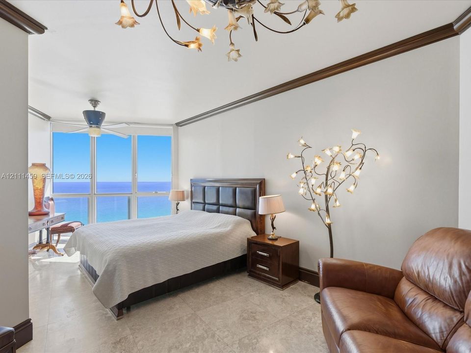 Floor-to-ceiling windows featuring ocean horizon, neutral stone flooring, decorative crown molding, and intricate multi-light chandelier