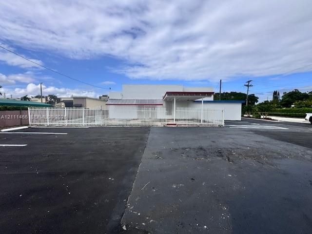 Commercial exterior featuring a paved parking lot, metal perimeter fencing, covered entryway, and white stucco facade