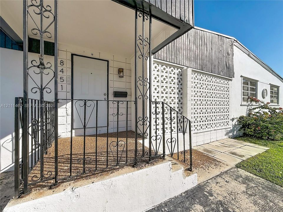 Mid-century modern entryway featuring decorative breeze block privacy screening, ornate wrought-iron railings, vertical wood-plank siding, and a concrete porch with aggregate surface texture