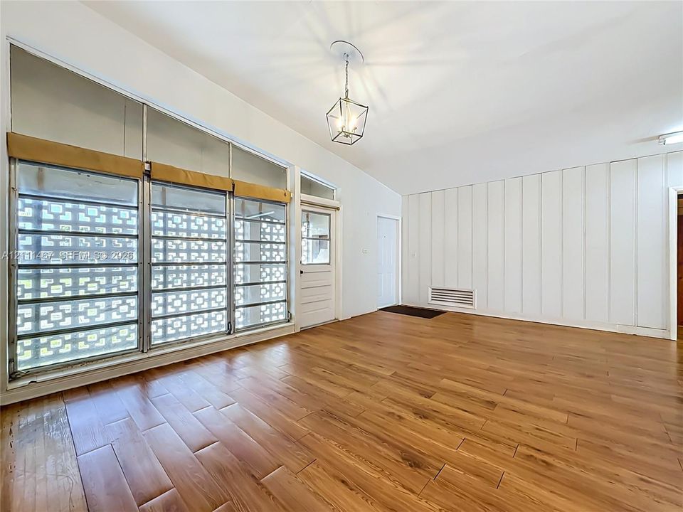 Living area featuring wood-finish flooring, vaulted ceilings, vertical wall paneling, a geometric lantern-style pendant light, and large windows with decorative breeze block screens