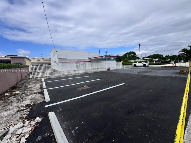 Asphalt-paved parking area featuring marked spaces, concrete parking bumpers, a perimeter metal fence, and an exterior structure with a covered entrance