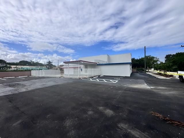 Commercial structure featuring a white exterior, expansive asphalt parking area, covered entrance, and perimeter fencing