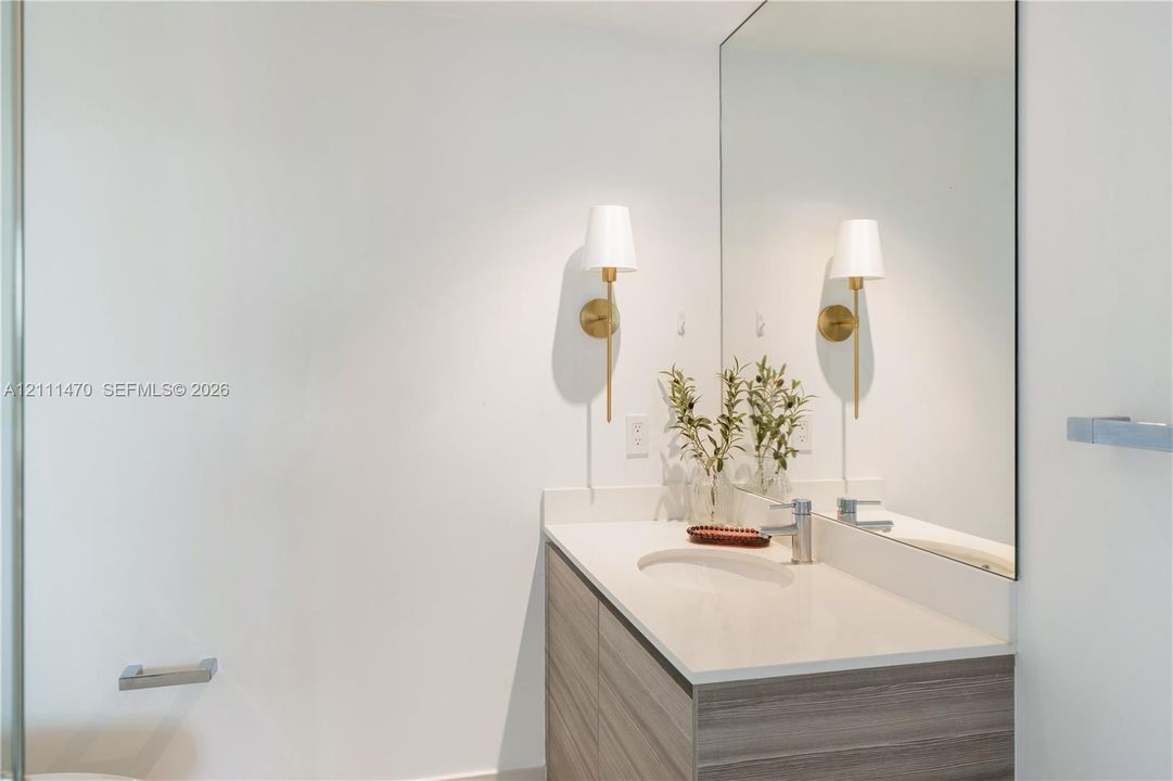 Modern vanity featuring a stone-composite countertop with an integrated oval sink, horizontal wood-finish cabinetry, a large frameless wall mirror, and dual brass-toned wall sconces