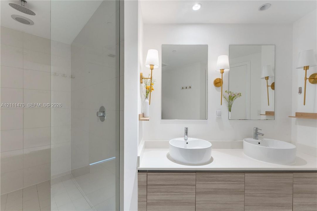 Dual-sink vanity with wood-finish cabinetry, rectangular vessel sinks, frameless wall mirrors, brass-toned wall sconces, and a glass-enclosed shower featuring a rain-style showerhead