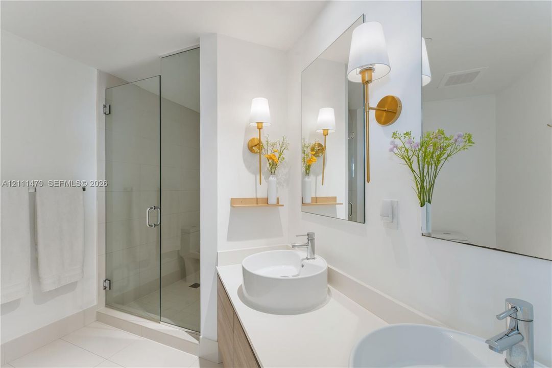 Bathroom featuring a dual-sink vanity with light-tone wood cabinetry, a quartz countertop, glass-enclosed shower, and gold-finish wall sconces