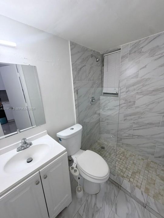 Bathroom featuring a walk-in shower with large-format wall tile and stone-finish floor tile, glass shower enclosure, white vanity cabinet with integrated sink, and wall-mounted vanity mirror