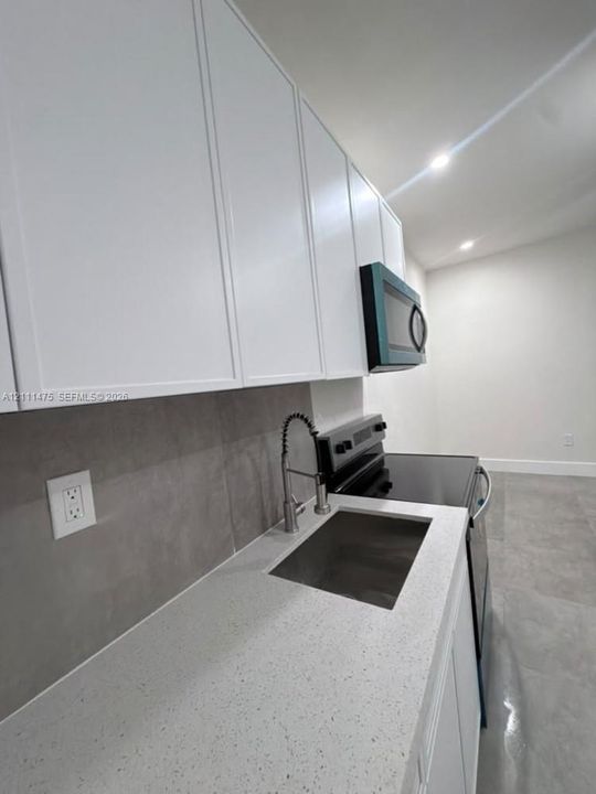 Shaker-style white cabinetry, solid surface countertops with integrated sink, large-format grey tile backsplash, stainless steel spring-neck faucet, and recessed ceiling lighting