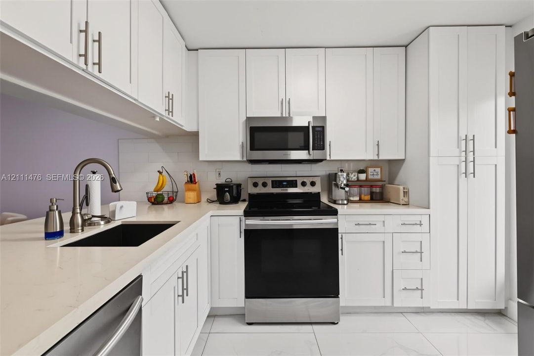 Kitchen featuring white shaker-style cabinetry, light-toned stone-surface countertops, subway tile backsplash, stainless steel appliances, and large-format tile flooring