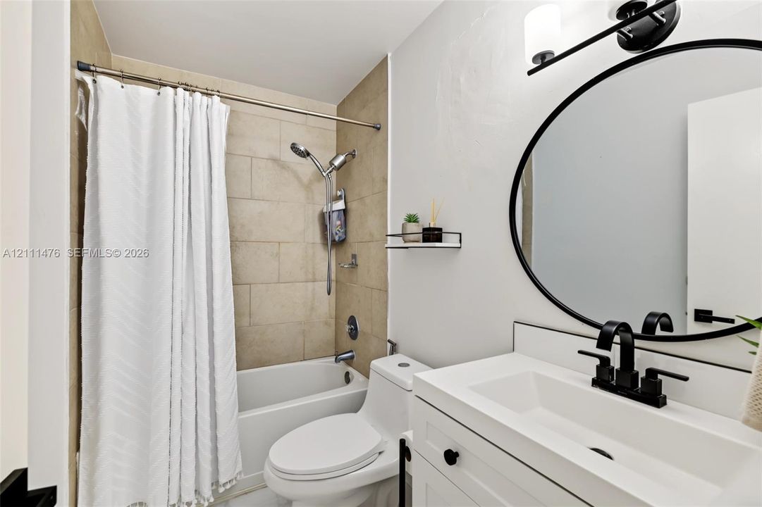 Bathroom featuring a white vanity with an integrated basin, matte black widespread faucet, circular mirror with a black frame, wall-mounted vanity lighting, and a bathtub with beige tiled surround