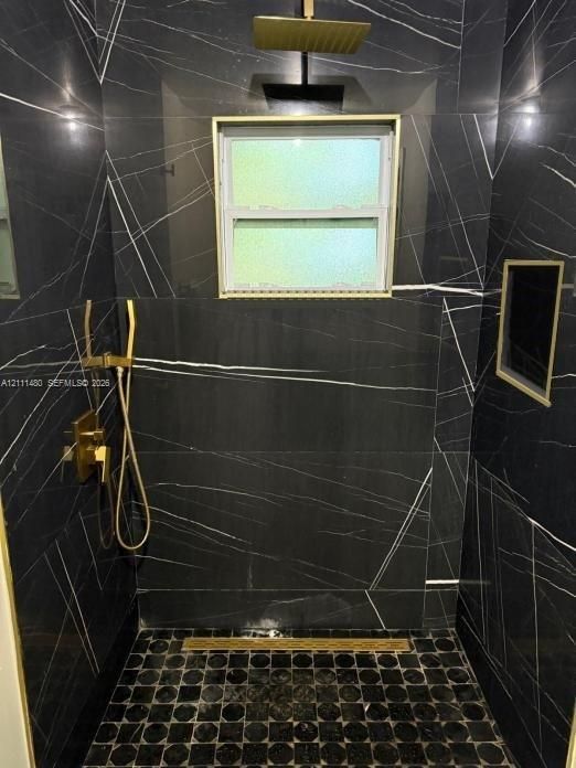 Shower enclosure featuring floor-to-ceiling black veined stone-finish tile, gold-tone rainfall shower head, handheld sprayer, recessed wall niche, and geometric tile floor inlay with a linear drain