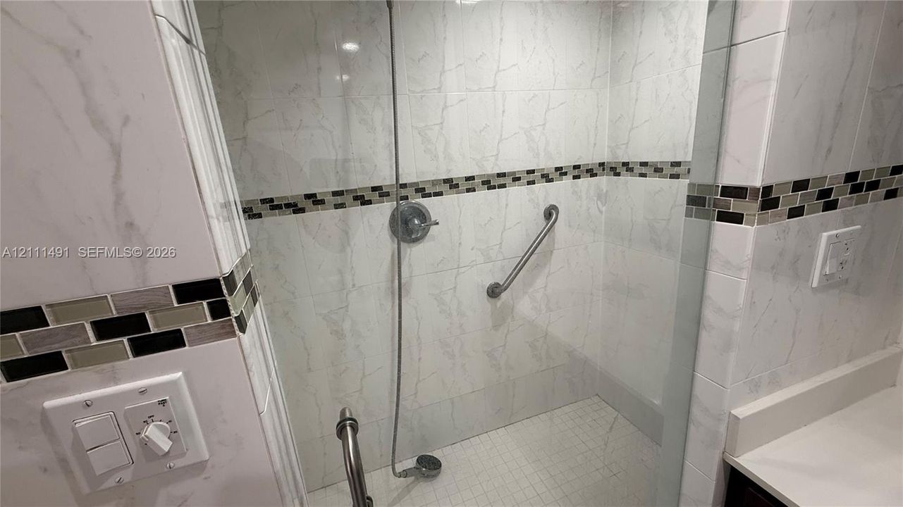 Walk-in shower enclosure featuring floor-to-ceiling large-format stone-patterned wall tile, decorative mosaic tile accents, a handheld shower fixture, and wall-mounted support bars