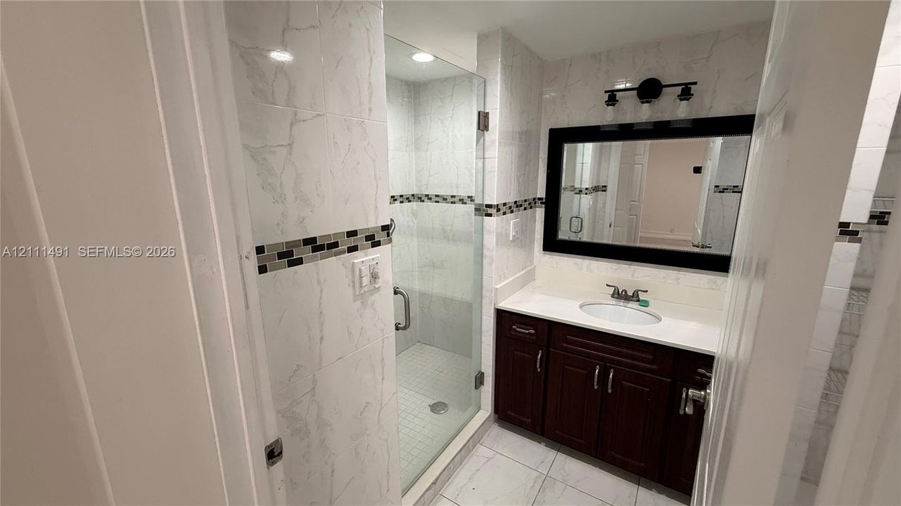 Bathroom featuring white stone-finish wall and floor tiling, dark wood-finish vanity with a white countertop, glass-enclosed walk-in shower, and a framed mirror with integrated lighting