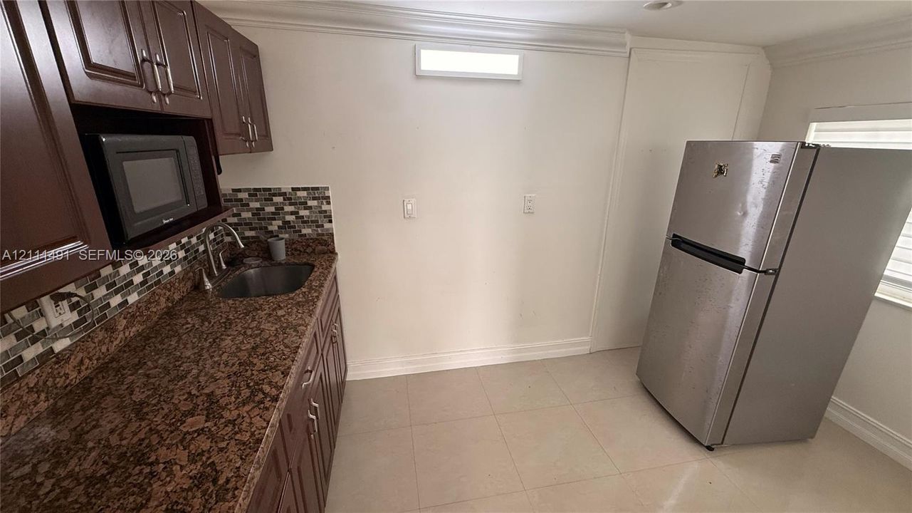 Kitchen area featuring dark-tone cabinetry, speckled stone-finish countertops, multi-toned glass tile backsplash, stainless steel sink with high-arc faucet, and tiled flooring