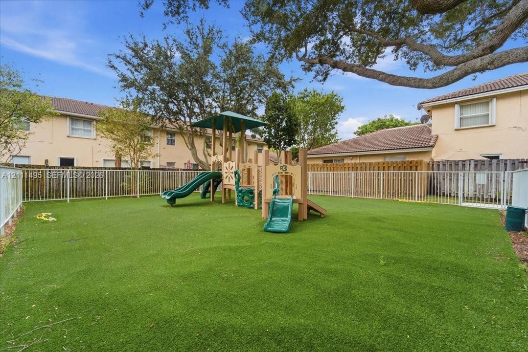 Common area featuring a synthetic grass lawn, perimeter metal fencing, and a permanent playground structure