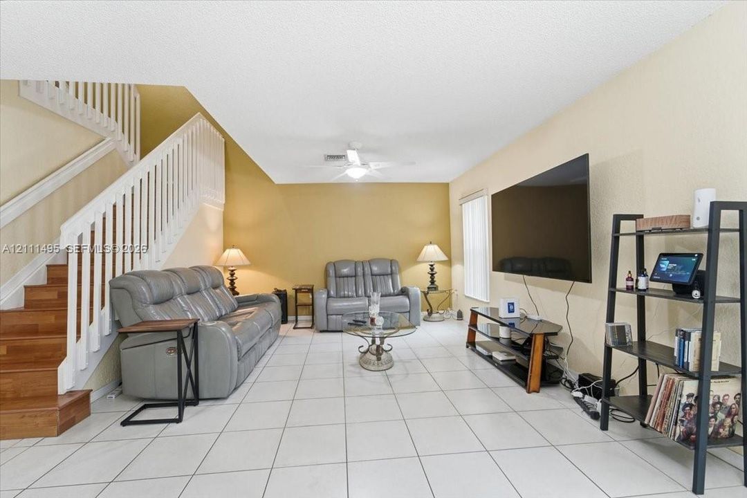 Living area featuring white ceramic tile flooring, a staircase with wood-finished treads and white balusters, and a ceiling-mounted light fixture with integrated fan