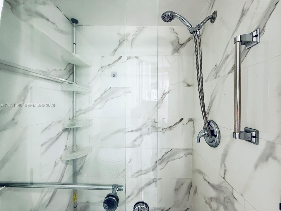 Shower stall featuring floor-to-ceiling stone-look tile, frameless glass door, wall-mounted handheld shower fixture, and metal grab bars