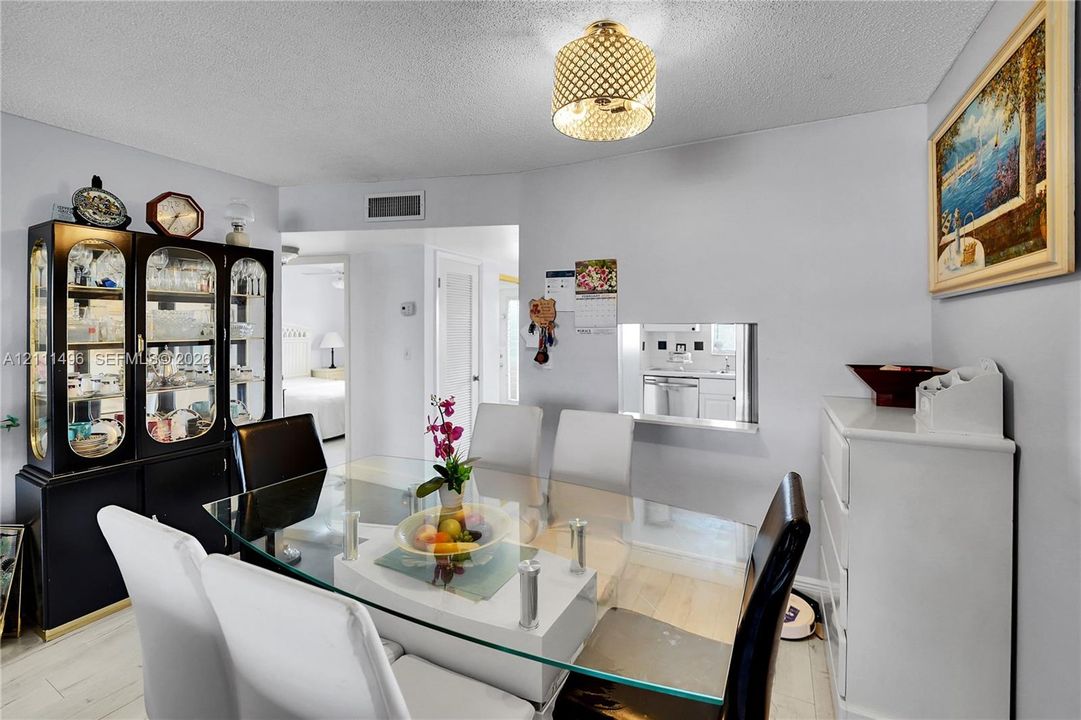 Dining area featuring light-toned wood-finish flooring, a contemporary cylindrical pendant light with decorative mesh detailing, and a wall cutout providing a glimpse into the kitchen area