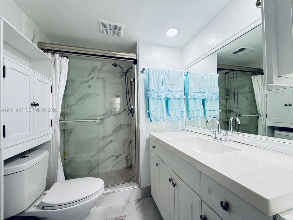 Bathroom featuring a shower with stone-patterned wall tile, a wide vanity with a solid-surface integrated sink, white shaker-style cabinetry, and a full-width wall mirror