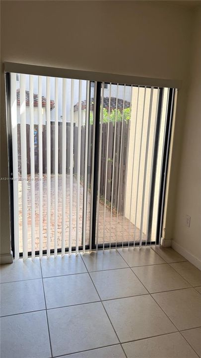 Sliding glass door with vertical blinds, neutral tile flooring, and a paved outdoor patio area enclosed by a wooden fence
