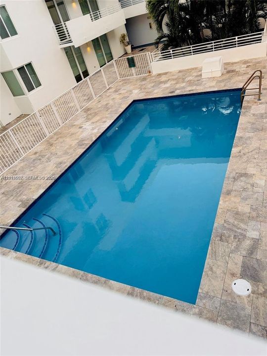 Rectangular swimming pool featuring stone-finish decking, integrated entry steps, metal handrails, and surrounding white metal perimeter fencing