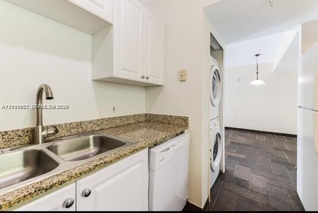 Kitchen area featuring stone-finish countertops, dual-basin stainless steel sink with high-arc faucet, white shaker-style cabinetry, built-in dishwasher, and a stacked laundry unit