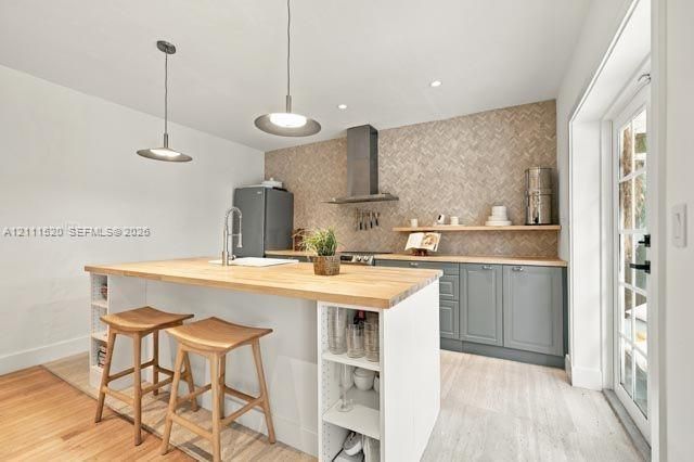 Kitchen featuring a wood-topped island with open shelving, herringbone-patterned tile backsplash, stainless steel range hood, grey shaker-style cabinetry, and floating wood shelving