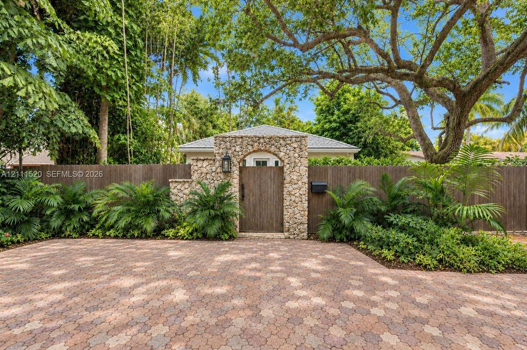 Paver driveway featuring a stone-clad arched entryway with a wood-finished privacy gate, surrounded by mature trees and lush landscaping