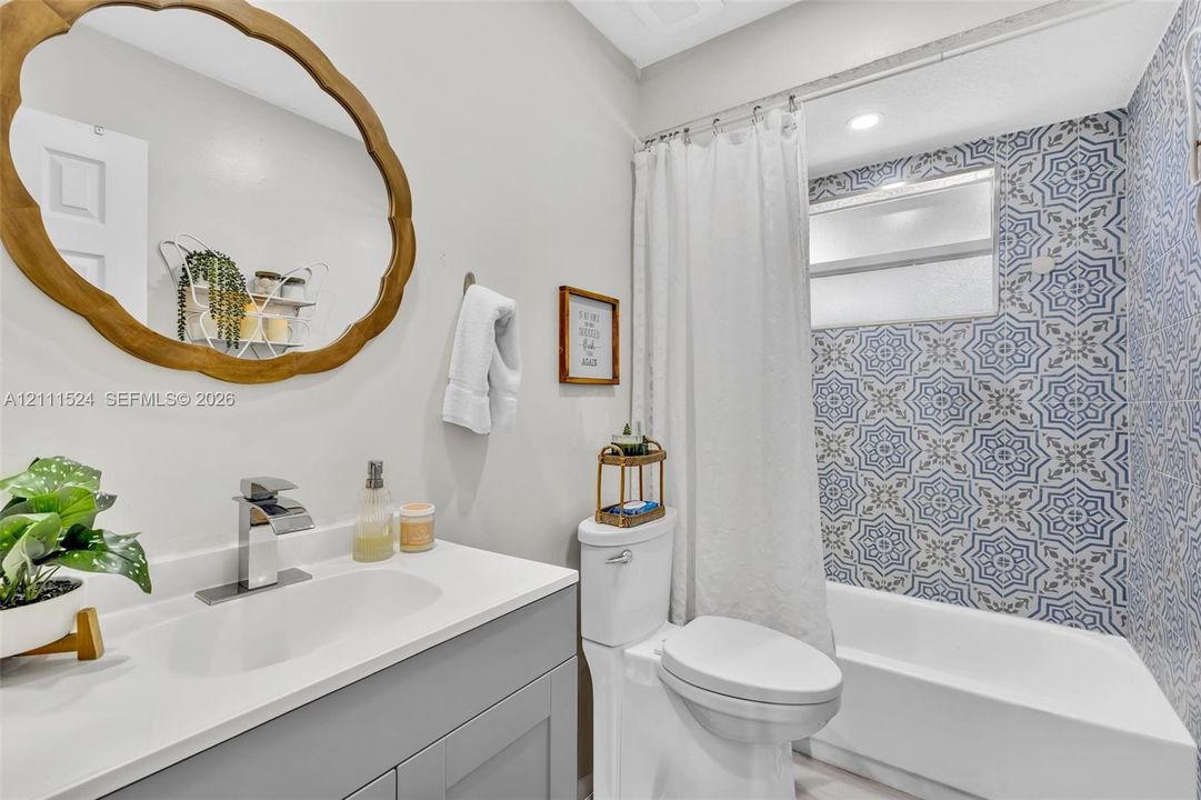 Gray vanity with a solid-surface countertop, integrated basin, chrome faucet, wall-mounted oval scalloped-edge mirror, and patterned tile shower surround