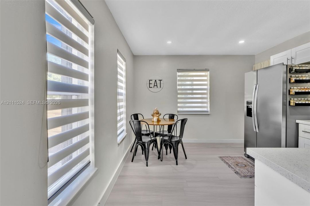Light-filled area featuring wood-finish flooring, dual-tone layered window shades, recessed lighting, stainless steel refrigerator, and crisp white cabinetry