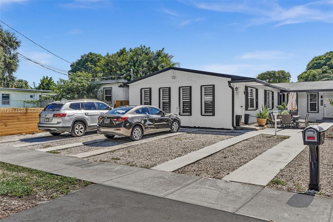 Single-story exterior featuring a light-toned vertical siding, dark trim accents, multiple windows with grid detailing, and a multi-car gravel driveway with concrete runners