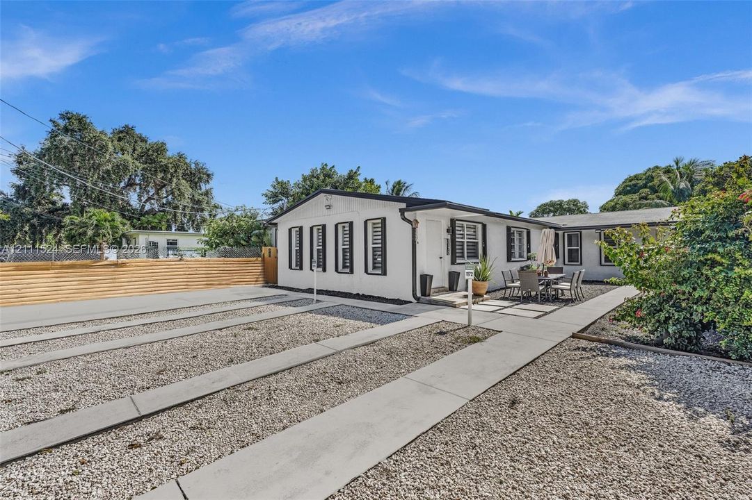 Single-story exterior featuring white stucco siding, contrasting dark window trim, a gravel-surfaced grounds area, concrete pathways, and a horizontal wood-slat perimeter fence