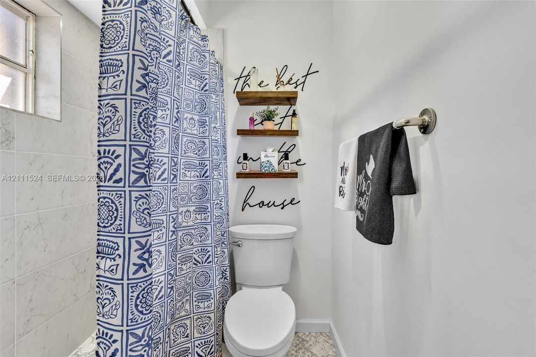 Three-tier wood wall shelving, wall-mounted metallic towel bar, light-toned stone-finish wall tile, and patterned tile flooring