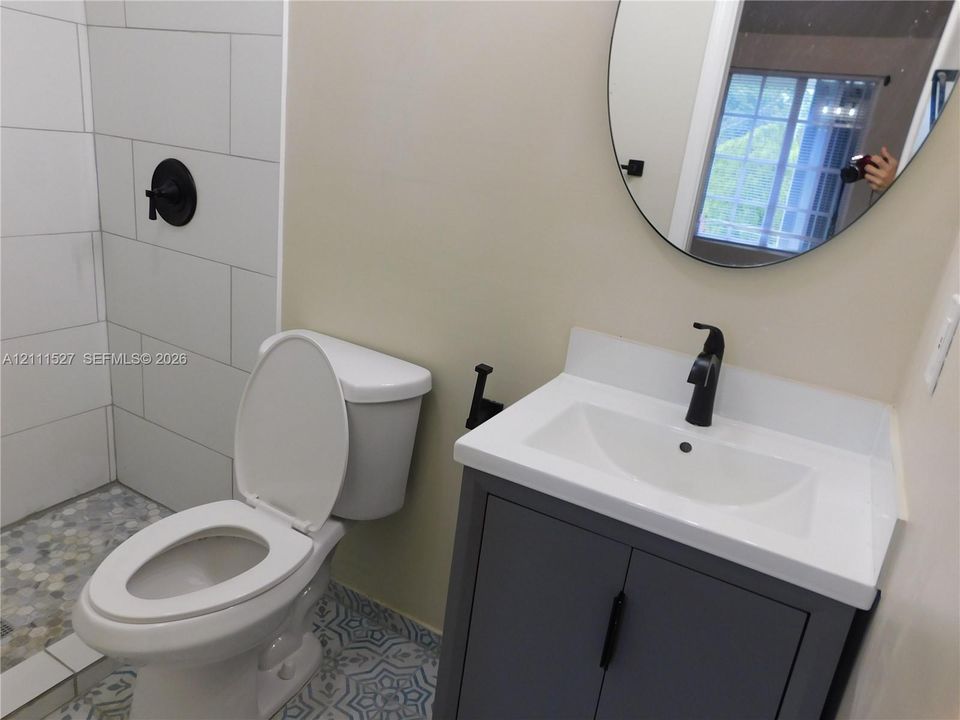 Bathroom featuring a gray vanity with a white countertop, matte black faucet, oval wall-mounted mirror, and a walk-in shower with large-format white tile and stone-texture floor tiling. Master bath