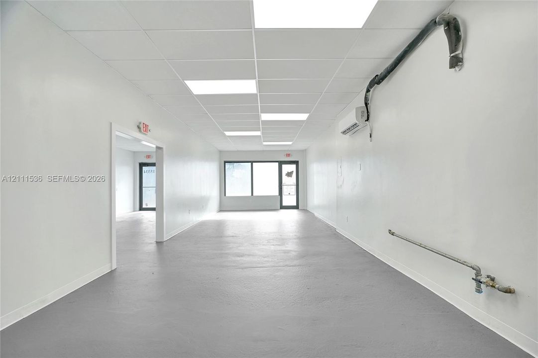 Open-concept interior featuring smooth wall finishes, a durable painted floor surface, drop ceiling with recessed lighting, wall-mounted climate control unit, and large aluminum-frame windows