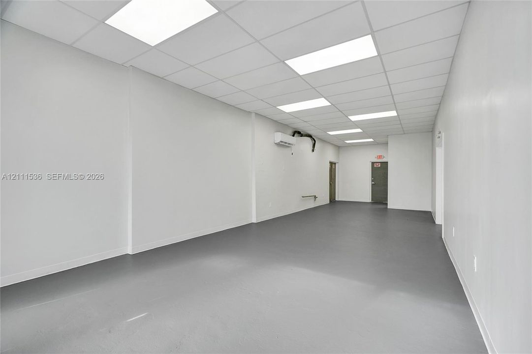 Open-plan interior featuring a drop ceiling with recessed panel lighting, smooth wall finishes, durable grey floor surfacing, and a wall-mounted climate control unit