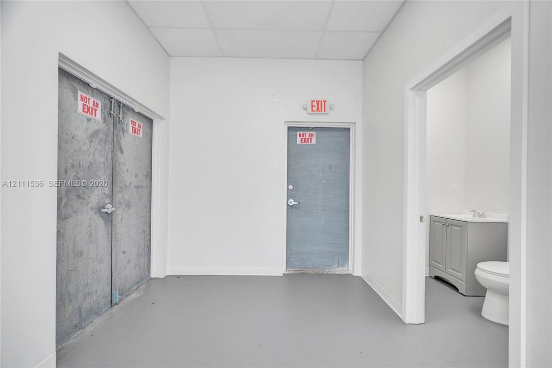 Neutral-toned interior space featuring double metal security doors, a secondary metal entrance, and a separate restroom area equipped with a vanity and commode