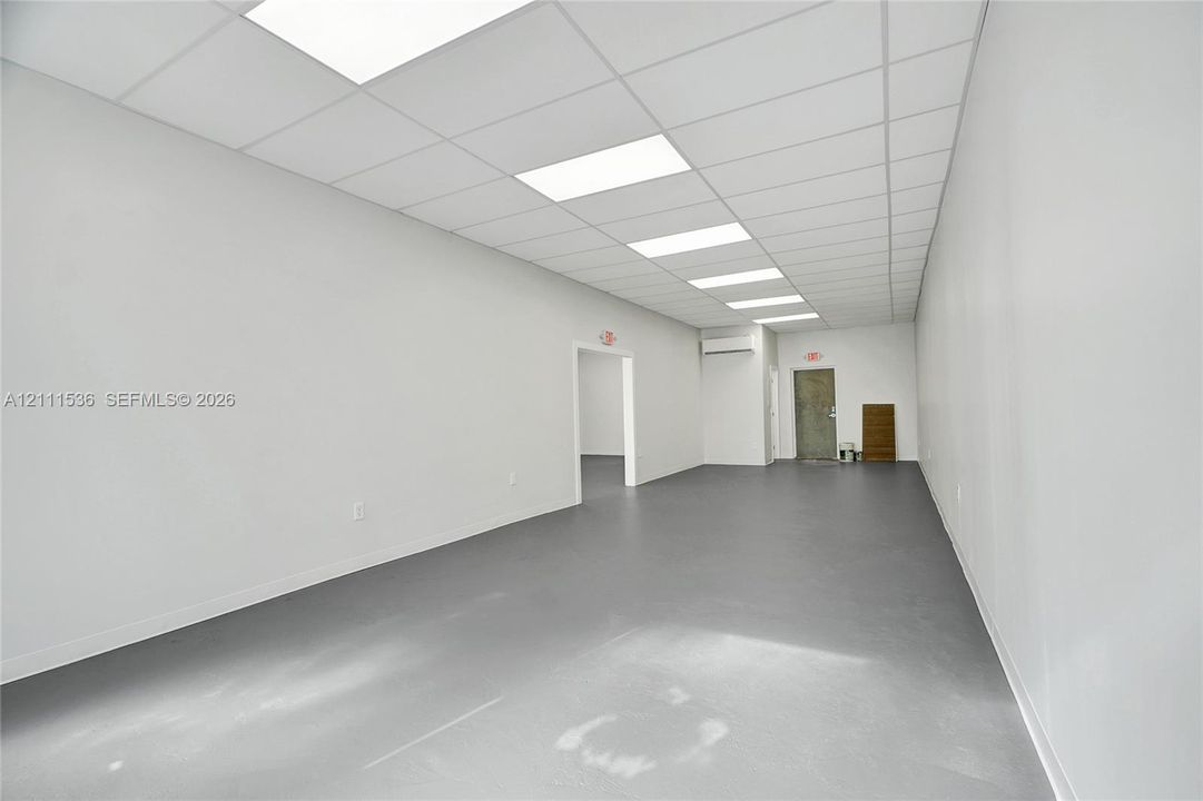 Expansive interior space featuring a drop ceiling with recessed rectangular lighting, smooth white walls, seamless grey flooring, and a wall-mounted ductless climate control unit
