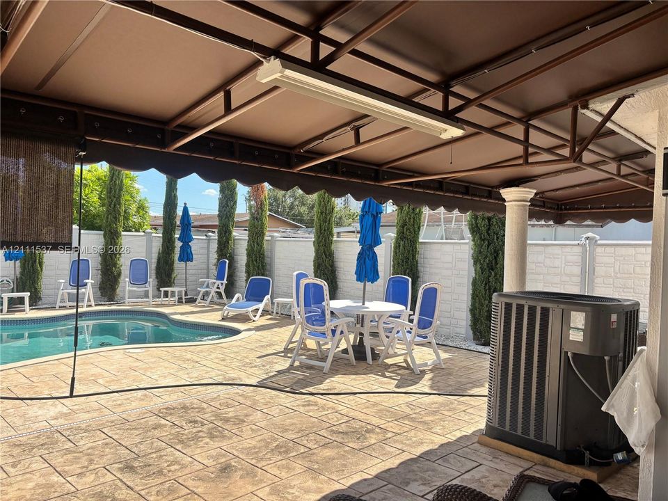 Covered patio with a metal frame and overhead lighting, stamped concrete surfacing, in-ground swimming pool, masonry privacy wall, and mature columnar evergreen trees