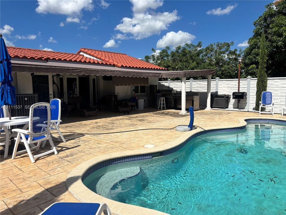 Outdoor living area featuring a custom swimming pool, stamped concrete patio, attached exterior covered awning, built-in outdoor kitchen, and perimeter privacy wall